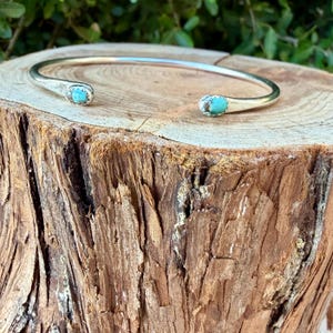 May include: A silver cuff bracelet featuring a smooth, polished metal band with an open design. Each end of the bracelet is capped with a small, round, bright blue turquoise stone set securely into the silver metal frame.