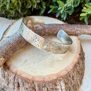 May include: A silver-toned metal cuff bracelet featuring an intricate embossed pattern of swirling scrolls, small stars, and circular motifs. The interior of the band is smooth and displays the stamped letters AK and 925 on the polished surface.