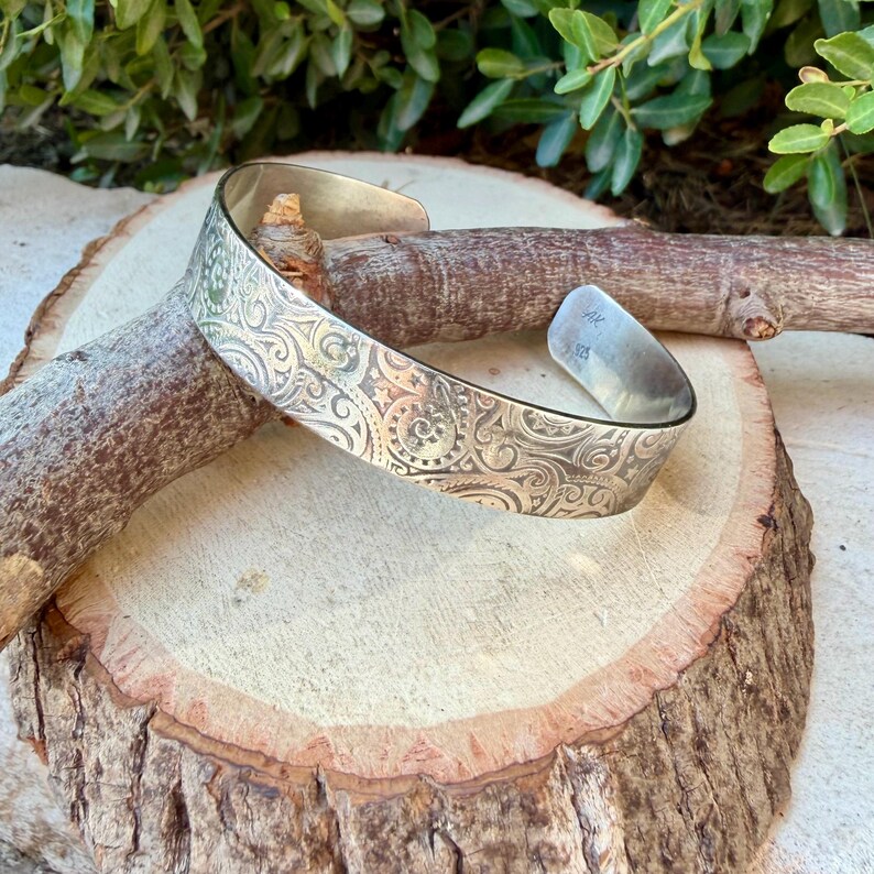 May include: A silver-toned metal cuff bracelet featuring an intricate embossed pattern of swirling scrolls, small stars, and circular motifs. The interior of the band is smooth and displays the stamped letters AK and 925 on the polished surface.