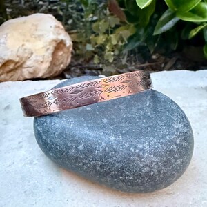 May include: A copper-colored metal cuff bracelet featuring a repeating engraved pattern of geometric shapes, including diamonds, chevrons, and horizontal lines. The bracelet has a polished finish and a rigid, open-ended circular band design.