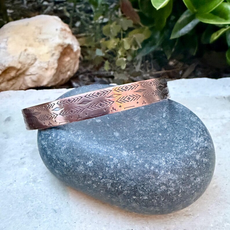 May include: A copper-colored metal cuff bracelet featuring a repeating engraved pattern of geometric shapes, including diamonds, chevrons, and horizontal lines. The bracelet has a polished finish and a rigid, open-ended circular band design.