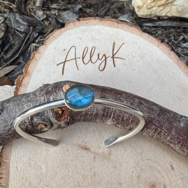 May include: A silver cuff bracelet with a blue gemstone set in the center. The bracelet has a simple, elegant design. The text "Allyk" is written on the wooden surface in the background.