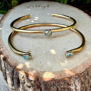 May include: Two gold-colored metal cuff bracelets. One bracelet has a single turquoise stone in the center, while the other has two turquoise stones, one at each end. The bracelets are displayed on a wooden surface with the handwritten word "Allyk".