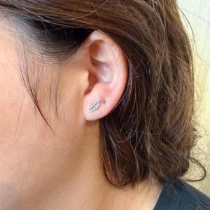 May include: A close-up of a silver feather stud earring on an ear. The earring is small and detailed, with a delicate feather design. The ear is visible against a blurred background.