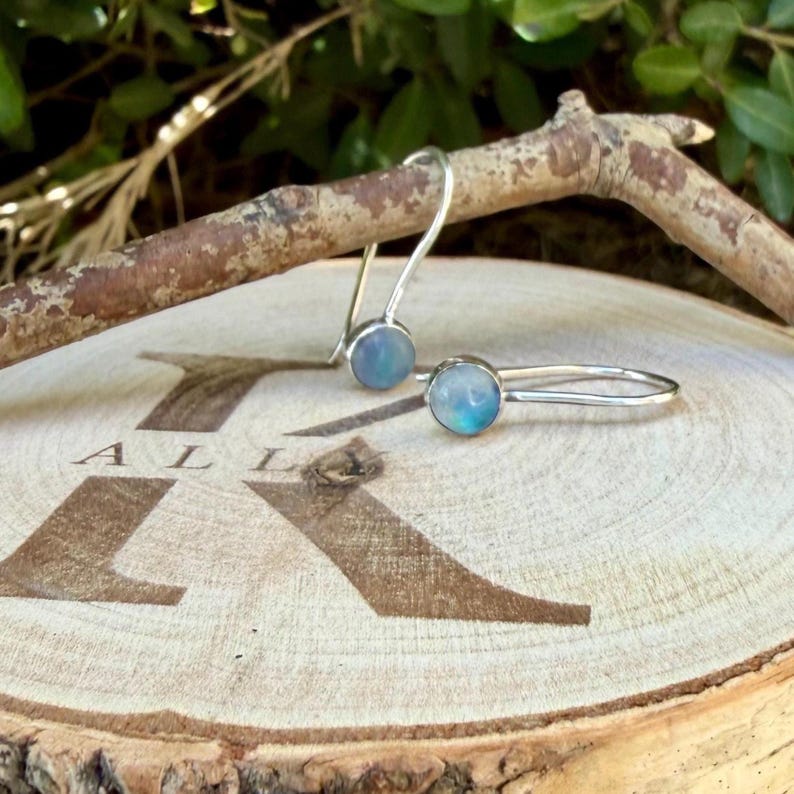 May include: A pair of silver hook earrings featuring round, iridescent blue cabochon stones. The earrings are resting on a circular wooden slice that has the letter K branded into the surface, positioned beneath a curved wooden branch.