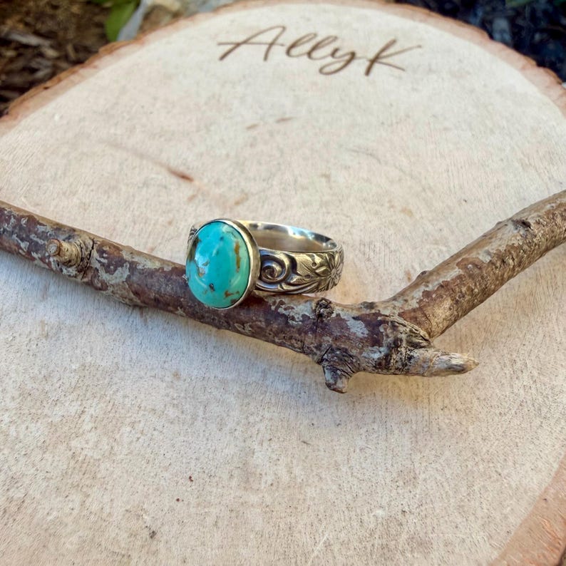 May include: A silver ring with a large, oval turquoise stone. The ring band features a floral design. The ring is displayed on a wooden branch, with the name "Ally K" lightly etched into the wooden background.