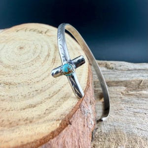 May include: A silver cuff bracelet featuring a cross design with a turquoise stone at the center. The cross is detailed with etched patterns. The bracelet is displayed on a wooden surface.