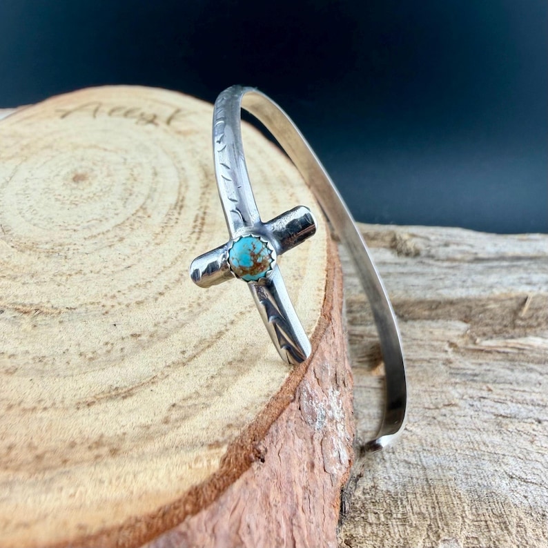 May include: A silver cuff bracelet featuring a cross design with a turquoise stone at the center. The cross is detailed with etched patterns. The bracelet is displayed on a wooden surface.