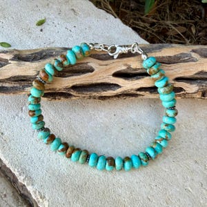 May include: A bracelet composed of round, turquoise-colored beads with brown matrix inclusions, interspersed with small, dark metallic spacer beads. The piece is secured with a silver-toned metal lobster claw clasp and a circular jump ring.