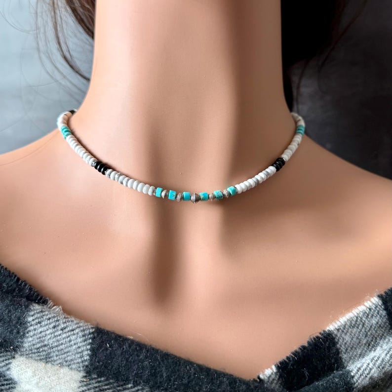 May include: A beaded choker necklace featuring white, black, and turquoise beads. The necklace is worn around the neck and is made of small, round beads. The necklace has a simple, minimalist design.
