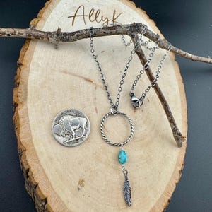 May include: A silver necklace with a turquoise stone and feather pendant. The necklace features a circular design and is displayed on a wooden surface with a silver coin. The word "Allyk" is written in cursive.