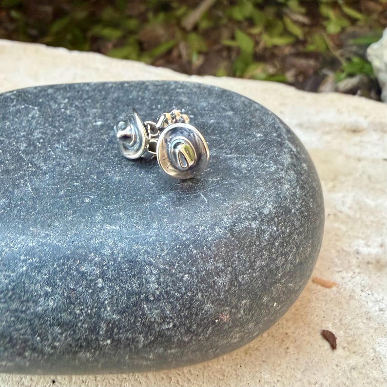 May include: A pair of silver-coloured cowboy hat earrings. The earrings are small, detailed, and have a classic cowboy hat shape. The earrings are displayed on a grey stone, with a blurred green and brown background.