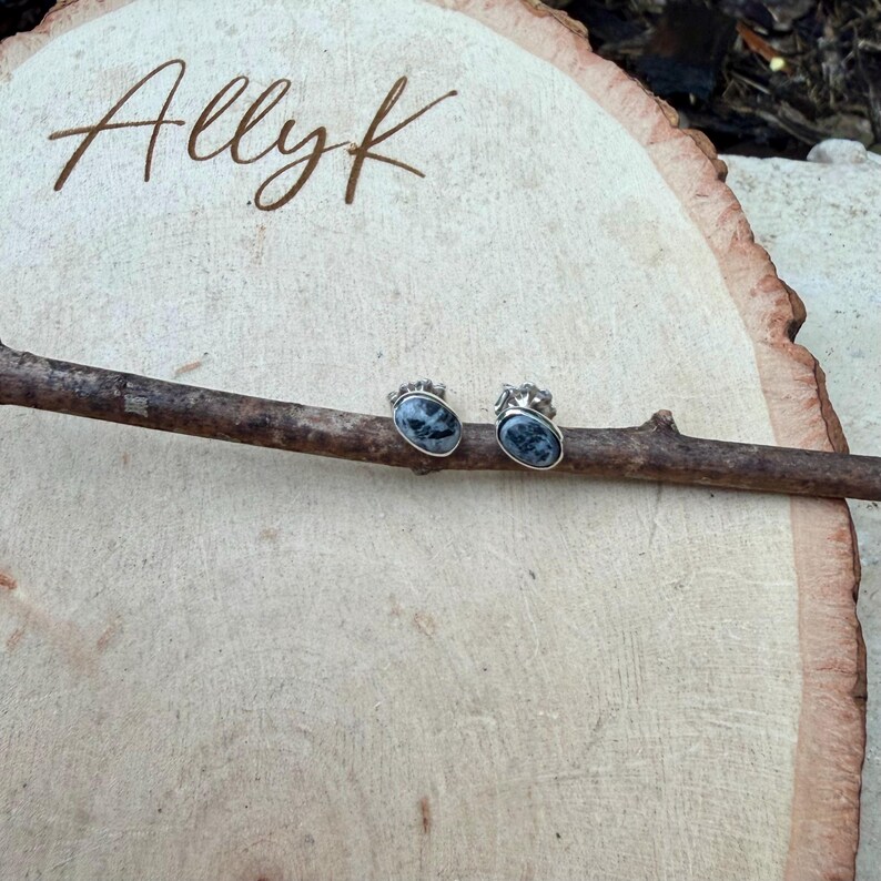 May include: A pair of oval-shaped stud earrings with a silver setting and a dark blue and gray stone. The earrings are displayed on a wooden branch, set against a light wood background. The word "Allyk" is written in cursive.