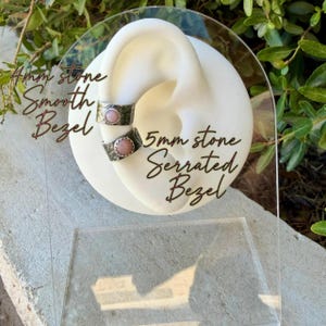 May include: Two silver-toned ear cuffs displayed on a white ear-shaped stand. The top cuff features a 4mm pink stone in a smooth bezel setting. The bottom cuff features a 5mm pink stone in a serrated bezel setting. Both cuffs have textured bands.