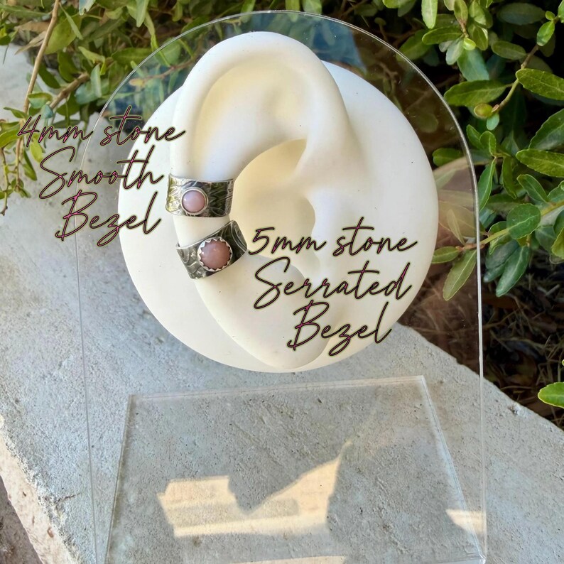May include: Two silver-toned ear cuffs displayed on a white ear-shaped stand. The top cuff features a 4mm pink stone in a smooth bezel setting. The bottom cuff features a 5mm pink stone in a serrated bezel setting. Both cuffs have textured bands.