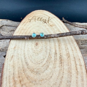May include: A pair of turquoise stud earrings with silver settings, displayed on a wooden plank with the engraved text "Ally K". The earrings are round and feature a bright blue-green stone.