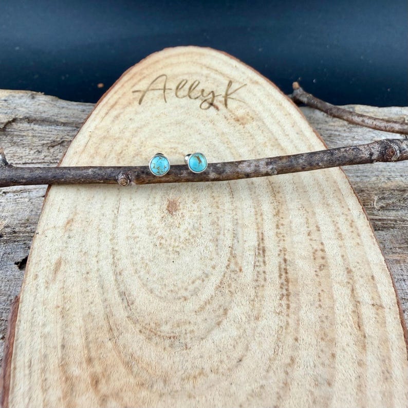 May include: A pair of turquoise stud earrings with silver settings, displayed on a wooden plank with the engraved text "Ally K". The earrings are round and feature a bright blue-green stone.