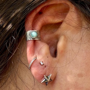 May include: A silver ear cuff featuring an engraved pattern and a round, light blue stone, worn alongside a thin silver hoop earring with small clear stones, a tiny rectangular stud, and a silver star-shaped stud earring on the earlobe.