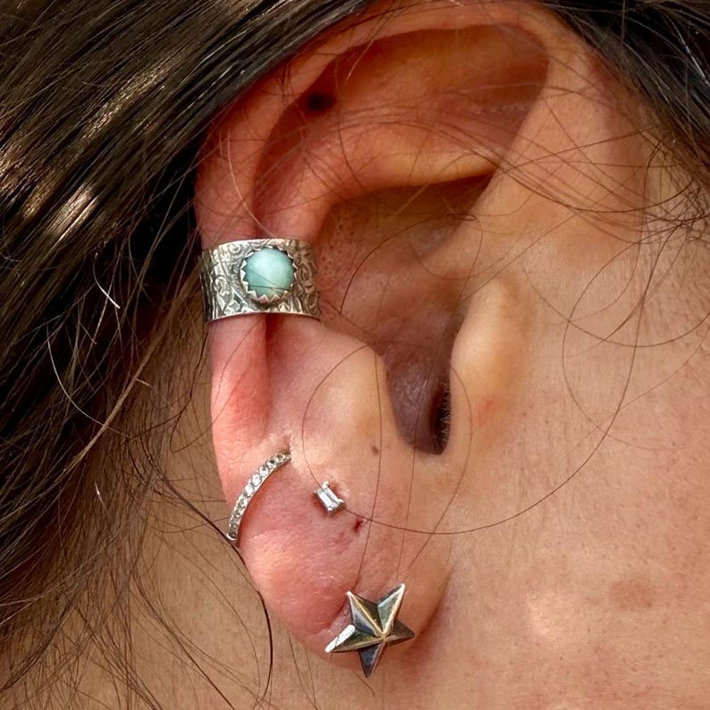 May include: A silver ear cuff featuring an engraved pattern and a round, light blue stone, worn alongside a thin silver hoop earring with small clear stones, a tiny rectangular stud, and a silver star-shaped stud earring on the earlobe.