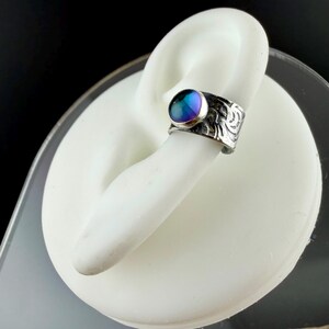 May include: A silver-toned ring with a large, iridescent, round gemstone. The ring has an ornate, etched design on the band. The gemstone displays a blend of blue, purple, and green hues. The ring is displayed on a white form.