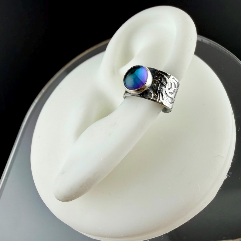 May include: A silver-toned ring with a large, iridescent, round gemstone. The ring has an ornate, etched design on the band. The gemstone displays a blend of blue, purple, and green hues. The ring is displayed on a white form.