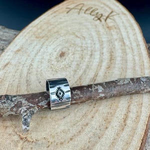 May include: A silver ring with a geometric design. The ring is wide and has a textured surface. The design is a diamond shape with lines inside. The ring is displayed on a wooden branch.