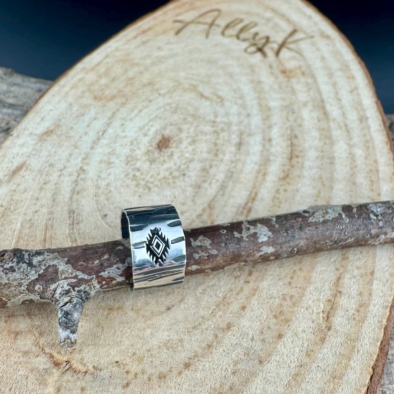 May include: A silver ring with a geometric design. The ring is wide and has a textured surface. The design is a diamond shape with lines inside. The ring is displayed on a wooden branch.