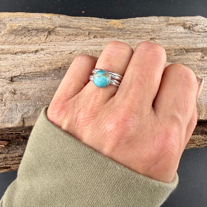 May include: A silver ring with a round, light blue turquoise stone. The ring has a braided band design and is worn on a finger. The image shows the ring on a hand, with a wooden background.
