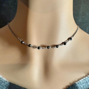 May include: A delicate necklace featuring small, dark beads strung along a silver-toned chain. The beads are evenly spaced, creating a minimalist and elegant design. The necklace is shown on a person's neck, highlighting its subtle beauty.