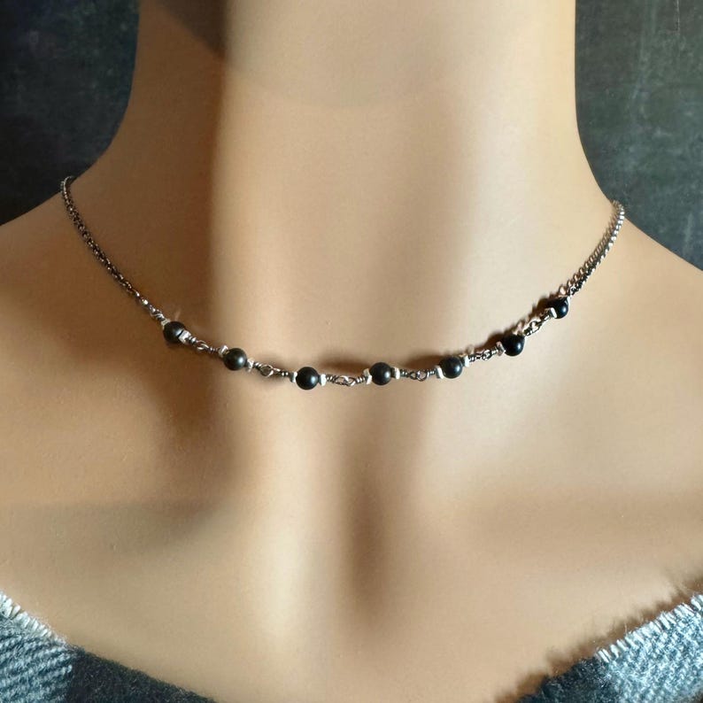 May include: A delicate necklace featuring small, dark beads strung along a silver-toned chain. The beads are evenly spaced, creating a minimalist and elegant design. The necklace is shown on a person's neck, highlighting its subtle beauty.