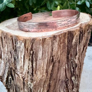 May include: A copper cuff bracelet featuring a repeating geometric pattern of etched diamonds, chevrons, and horizontal lines. The metal has a polished finish with darkened recessed areas that highlight the intricate stamped design details.