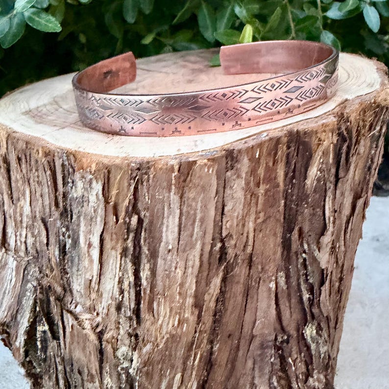 May include: A copper cuff bracelet featuring a repeating geometric pattern of etched diamonds, chevrons, and horizontal lines. The metal has a polished finish with darkened recessed areas that highlight the intricate stamped design details.