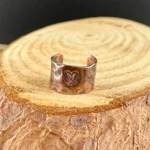 May include: A copper-colored ear cuff with a hammered texture and a butterfly design. The cuff is open-ended and sits on a wooden surface. The cuff is a jewelry item.