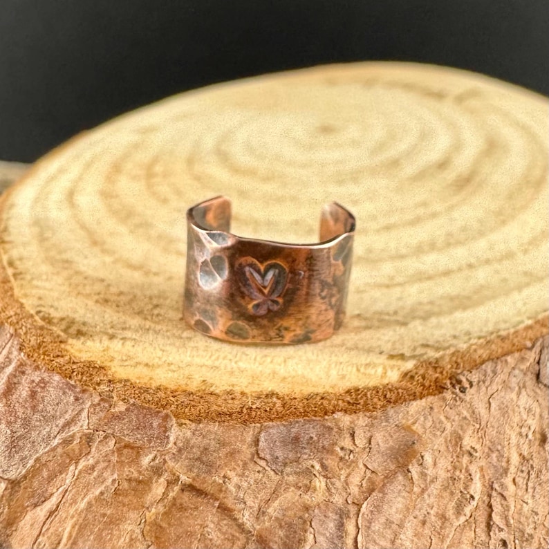 May include: A copper-colored ear cuff with a hammered texture and a butterfly design. The cuff is open-ended and sits on a wooden surface. The cuff is a jewelry item.