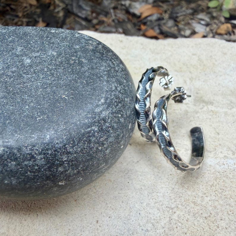 May include: A pair of silver hoop earrings with a textured, woven design. The earrings are crescent-shaped and have a post closure. The earrings are displayed next to a large, gray stone on a light-colored surface.
