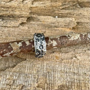 May include: A silver ring with a floral design. The ring is set against a wooden background. The ring's surface has a textured appearance, with detailed floral patterns.