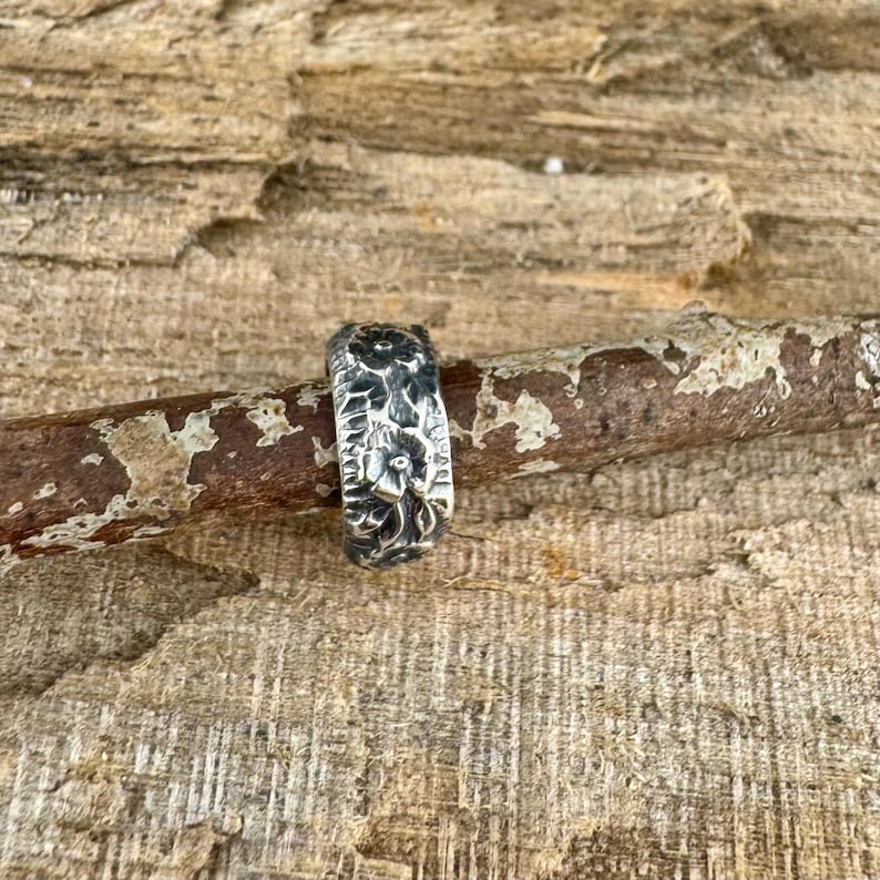 May include: A silver ring with a floral design. The ring is set against a wooden background. The ring's surface has a textured appearance, with detailed floral patterns.