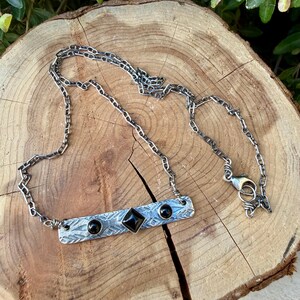 Pu&ograve; includere: Una collana in metallo color argento con un ciondolo a barra rettangolare caratterizzato da un motivo geometrico inciso. Il ciondolo &egrave; impreziosito da una pietra nera centrale a forma di diamante e due pietre nere rotonde ai lati.