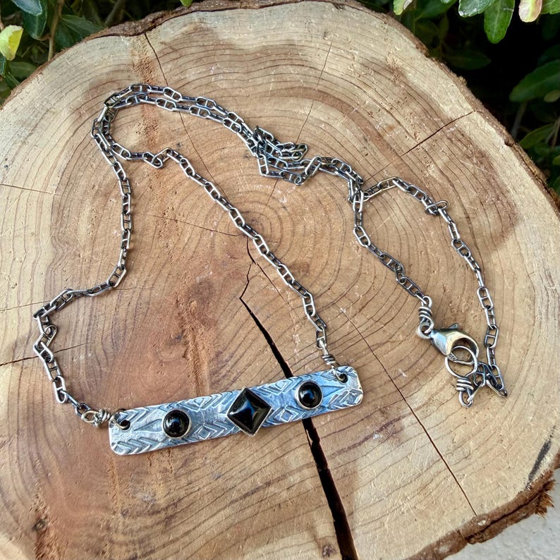 Pu&ograve; includere: Una collana in metallo color argento con un ciondolo a barra rettangolare caratterizzato da un motivo geometrico inciso. Il ciondolo &egrave; impreziosito da una pietra nera centrale a forma di diamante e due pietre nere rotonde ai lati.