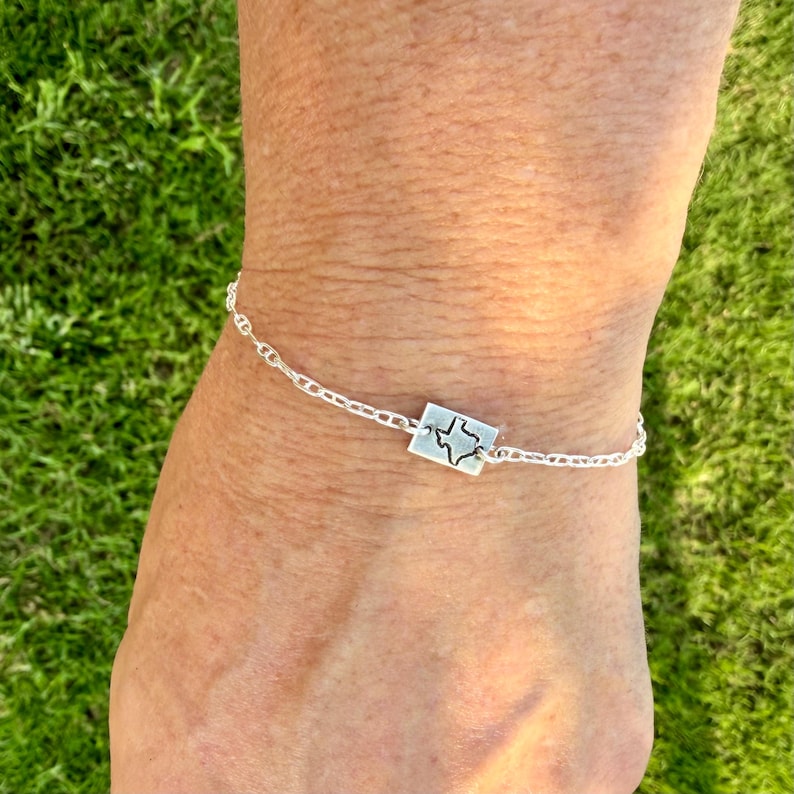 May include: A silver bracelet featuring a rectangular charm with the outline of the state of Texas. The bracelet is made of a delicate chain and is worn on a wrist against a backdrop of green grass.
