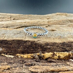 May include: A silver-toned ring with a twisted band design. The ring features a small, round gemstone with a light brown center and a turquoise border. The ring is set against a textured wooden surface.
