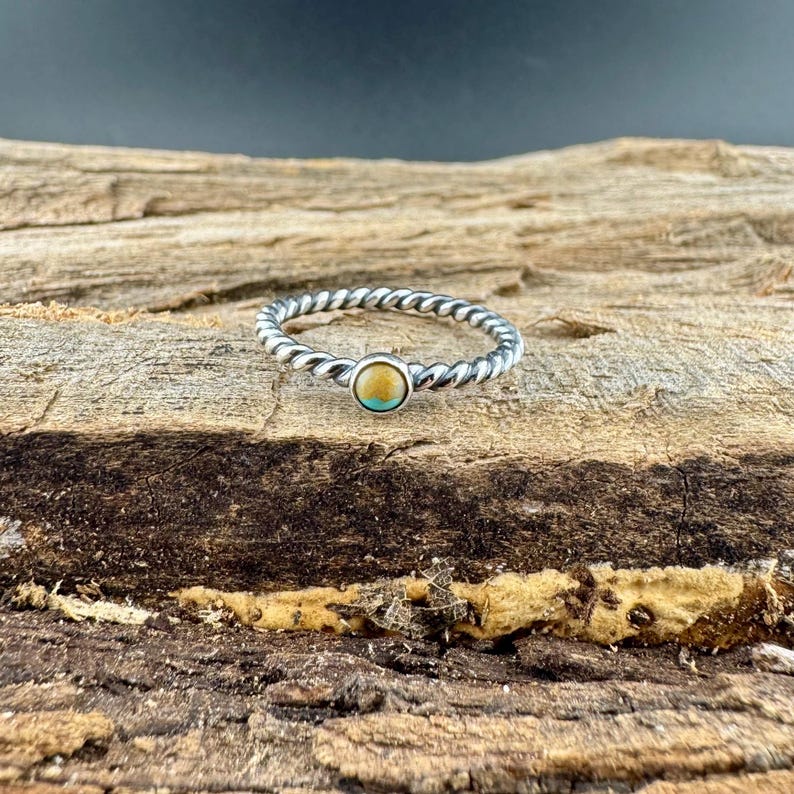 May include: A silver-toned ring with a twisted band design. The ring features a small, round gemstone with a light brown center and a turquoise border. The ring is set against a textured wooden surface.