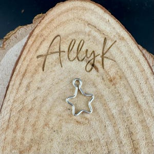 May include: A small, silver-colored star charm with a loop at the top. The charm is set on a light brown, wooden surface with the engraved text "Allyk". The wood has a natural grain and texture.