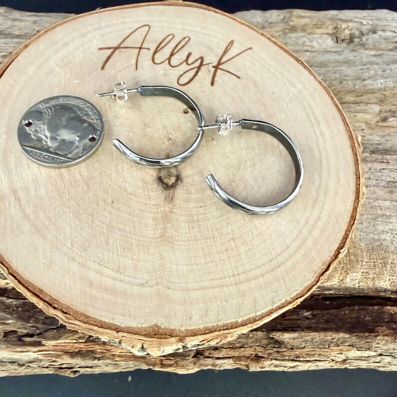 May include: Silver hoop earrings with a textured design, displayed on a wooden slice. A US nickel coin is next to the earrings for size comparison. The wooden slice has the name "Ally K" engraved on it.