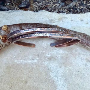 May include: Two copper cuff bracelets featuring a dark, recessed geometric pattern etched along the outer surface. The bracelets have an open-ended design with smooth, rounded edges and a polished metallic finish that reflects light across the surface.