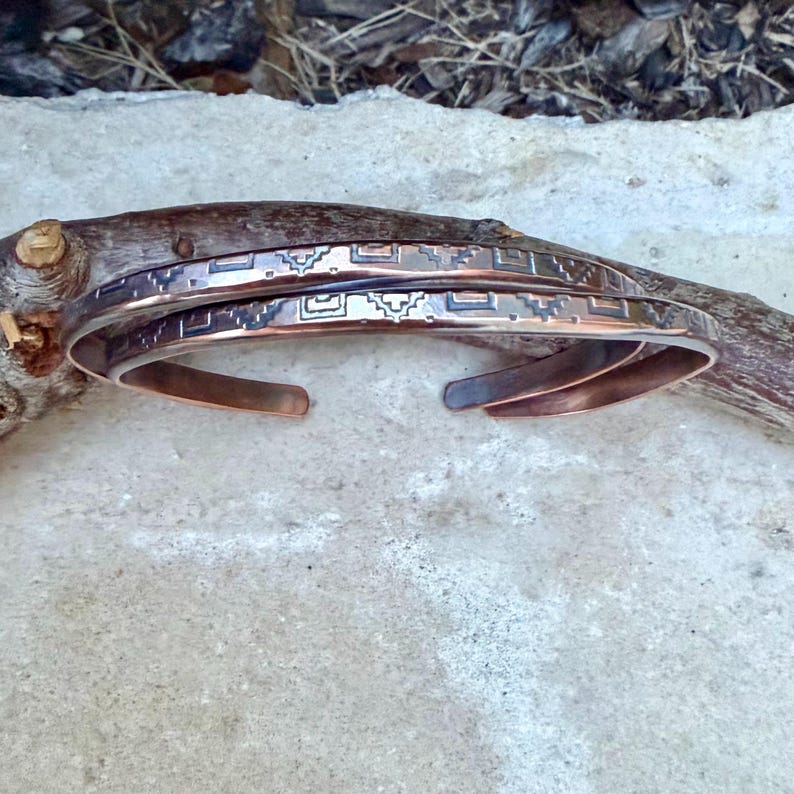 May include: Two copper cuff bracelets featuring a dark, recessed geometric pattern etched along the outer surface. The bracelets have an open-ended design with smooth, rounded edges and a polished metallic finish that reflects light across the surface.