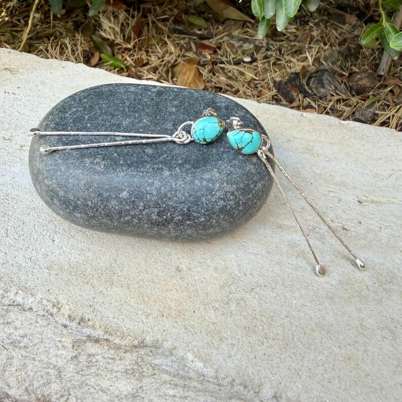 May include: A pair of long silver drop earrings featuring teardrop-shaped turquoise stones with dark matrix veining. Each earring consists of a thin, textured silver wire stem that extends downward from the stone, ending in a small silver bead.