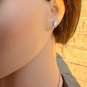 May include: A close-up of a person's ear with a small, silver arrowhead stud earring. The earring is detailed with a textured surface. The background is blurred, with warm tones of yellow and brown.