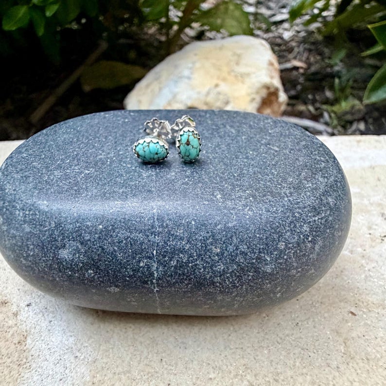 May include: A pair of oval turquoise stud earrings with silver-colored settings. The earrings are displayed on a smooth, gray stone, showcasing their vibrant blue-green color. The earrings are a classic jewelry item.