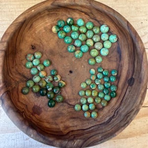 May include: A round, dark brown wooden bowl containing three distinct clusters of small, round, turquoise-colored beads. The beads exhibit natural variations in color, ranging from light blue-green to deep forest green, with some showing brown matrix patterns.